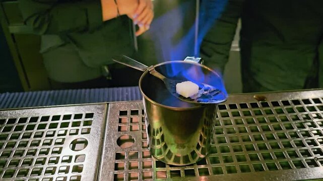 Close-up of traditional absinthe preparation at a dim cocktail bar, with blue flame, sugar cube on a slotted spoon, metal cup, cut glass goblet, and stainless counter for nightlife videos.
