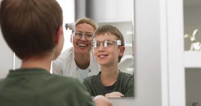 Mirror reflection of happy child trying on first prescription glasses at optical store enjoying stylish look and supportive female optometrist assisting with frame selection evaluating fit and comfort