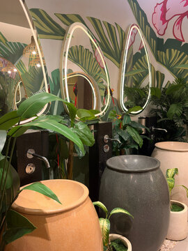 A modern restroom features several decorative planters in earthy tones. Tall green plants and stylish mirrors create a fresh, inviting atmosphere with tropical motifs.