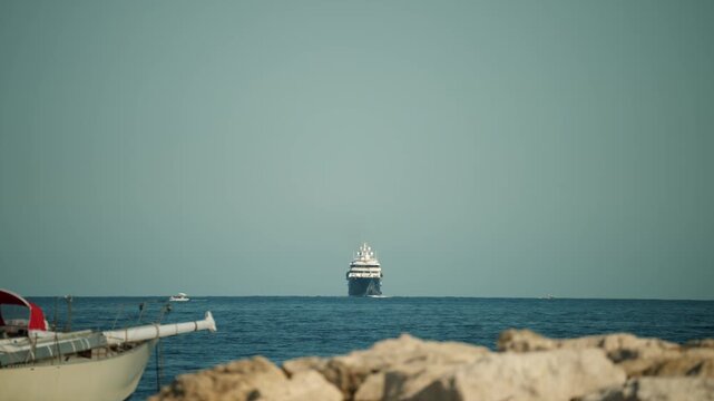Luxury yacht cruising on the vast blue ocean with a clear sky, rocks and sailboat in the foreground. Travel and nautical theme
