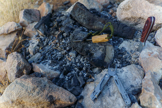 Ferro rod, multitool, and knife resting on campfire ashes with charred wood and stones, highlighting outdoor readiness and survival skills