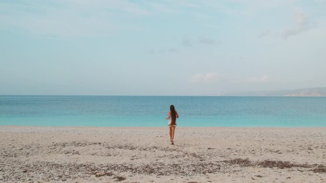 Woman heading toward turquoise sea on wide pebble beach under soft morning sky