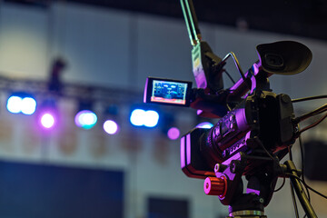 A professional video camera recording a live stage event with colorful bokeh lights in the...
