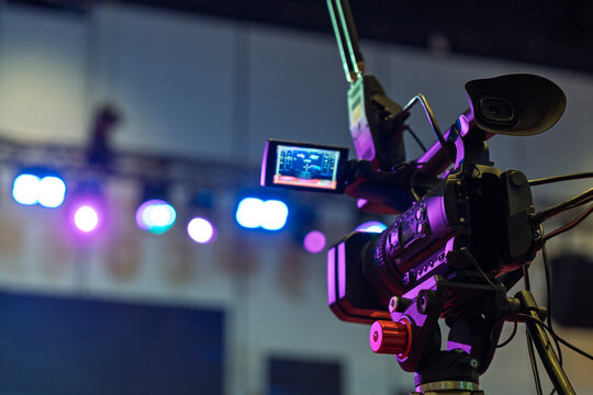 A professional video camera recording a live stage event with colorful bokeh lights in the background.