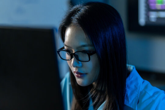 Close-up of an Asian woman developer in glasses looking down in concentration, lit by cool blue light with a code screen blurred behind her.