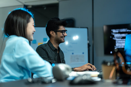 Asian woman and South Asian man in glasses smile and laugh while working at a desk of code in a dark blue tech office at night.