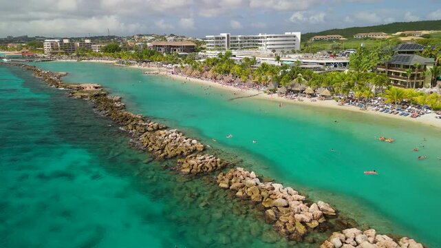 Aerial orbit shot of Mambo Beach in Curacao revealing turquoise Caribbean water, rocky breakwater, thatched umbrellas, crowded sandy shore and resort hotels