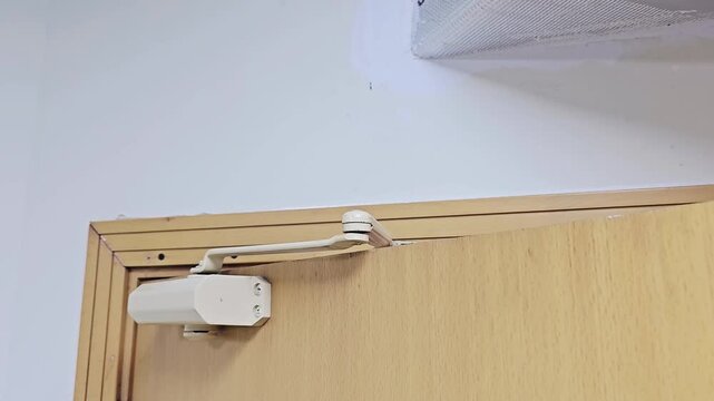 An automatic hydraulic door closer is mounted on a light wooden door, showing the mechanical arm mechanism.