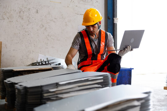 Construction worker in hard hat and high visibility vest uses a laptop beside stacked metal sheets inside an industrial warehouse.