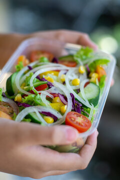 Fresh vegetable salad with lettuce, cucumber, tomato, corn and onion in takeaway box held by hands. Healthy diet, clean eating, meal prep and lifestyle concept.