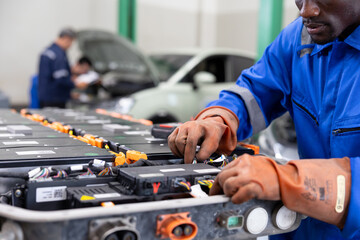 Technician wearing protective gloves services an electric vehicle battery pack in an auto workshop, inspecting modules and connections with tools beside a car. © akarawit