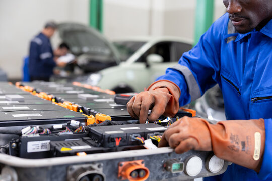 Technician wearing protective gloves services an electric vehicle battery pack in an auto workshop, inspecting modules and connections with tools beside a car.