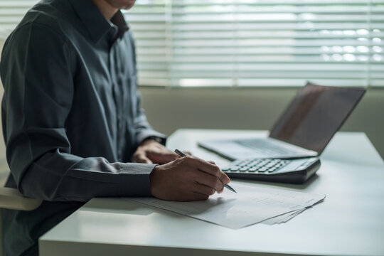 Close-up of businessman holding pen writing on financial document at office desk with calculator and laptop.