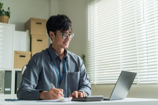 Young office worker wearing glasses smiling happily while writing on document and using laptop at desk.