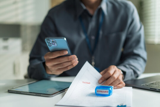Close-up of office worker using smartphone and checking approved document stamp for mobile transaction at desk.