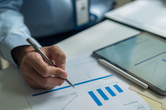 Close-up of businessman holding pen analyzing financial chart report at desk with digital tablet.