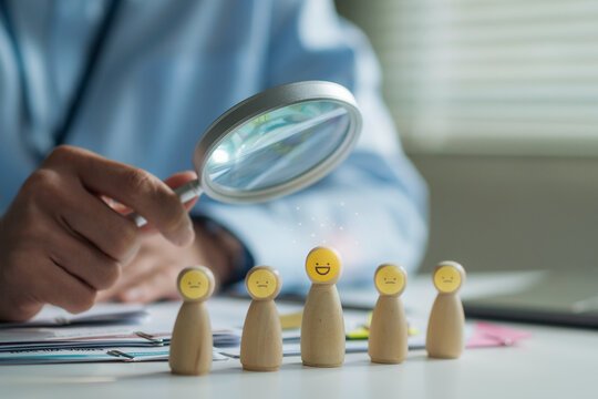 Close-up of HR manager using magnifying glass to select smiley wooden doll for recruitment and customer satisfaction concept.