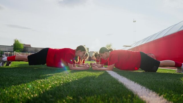 Womens soccer team core strength training on sunny artificial turf field, focused athletes in red jerseys holding plank position during pre-match conditioning