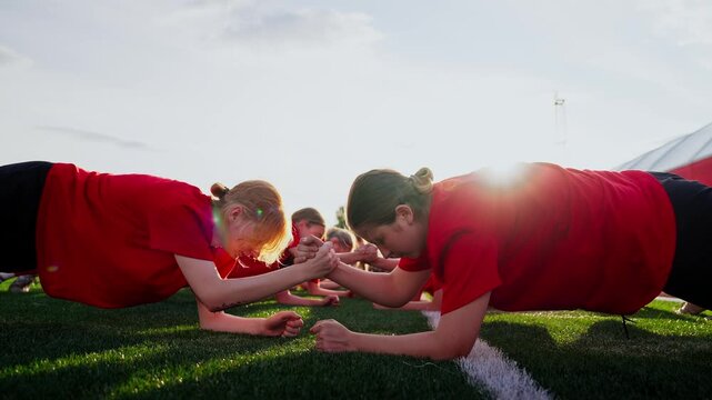 Focused womens soccer team outdoor training, teammates in red jerseys performing partner plank drills and strength exercises together on artificial turf at sunset