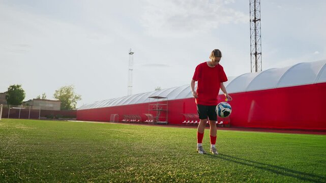 Female soccer player practicing ball control on green training field near red stadium wall during solo outdoor practice session at sunset for skill development
