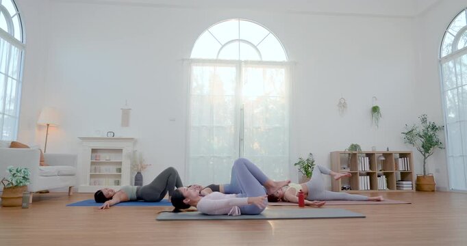 Asian adult female friends lie on yoga mats for supine spinal twist, moving knees side to side while trainer guides breathing, mobility, posture release, calm recovery routine inside bright studio