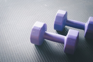 Obraz premium Close up of two purple dumbbells resting on a black textured gym mat with natural light. Home workout, strength training, weight loss, and active fitness lifestyle concept with copy space.