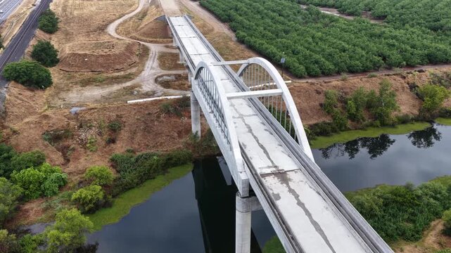 Arched concrete bridge spans quiet river near Fresno, California, USA, forming part of unfinished high-speed rail corridor, framed by dense orchards, reflective water. Aerial drone orbit shot.
