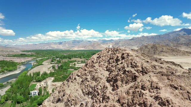 High-angle drone tracking and ascending shot rising over a rugged rocky ridge to reveal a white Buddhist chorten stupa overlooking the lush green Indus River valley in Leh Ladakh, India.