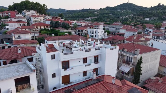 Drone circles around boutique resort property with rooftop terrace restaurant in old town square of Pefkochori surrounded by mediterranean pine tree mountains in background