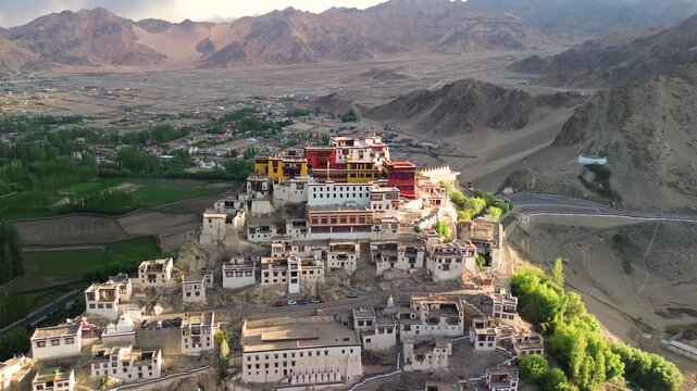 Drone tracking shot pulling back and rising over the historic tiered Thiksey Monastery complex during a warm sunset in Leh Ladakh, India.