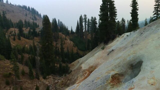 Tilt down view of colorful hydrothermal landscape and steaming fumarole on volcanic hillside in Lassen Volcanic National Park, California, surrounded by conifer forest and rugged mountain terrain, USA