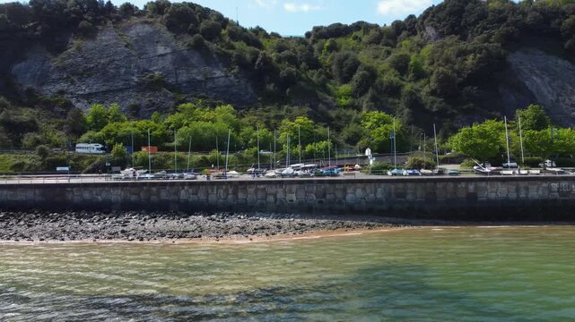 Aerial drone footage showing the rocky coastline, green cliffs, parked boats, and marina buildings at Mumbles in Wales on a clear day.