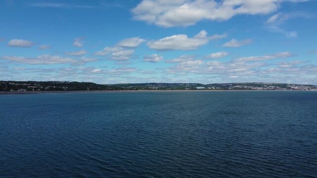 Calm blue sea water stretches under a bright sky with scattered clouds toward the distant Mumbles coastline in Wales, creating a peaceful coastal atmosphere.