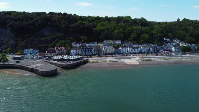 Aerial drone footage showing the coastline, beach, and colorful houses of Mumbles village with lush green hills in the background under clear blue sky.
