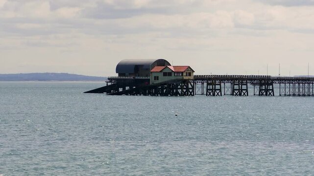 A tranquil view of a wooden pier and boathouse extending over the calm sea waters under a cloudy sky at Mumbles in Wales.