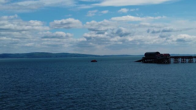 Calm blue sea stretches toward distant hills under a partly cloudy sky with Mumbles pier visible on the right side.