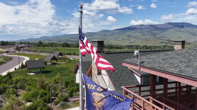 American flag flies beside state flag atop welcome center rooftop in rural Oregon, USA, overlooking green valley, winding road, distant mountains under partly cloudy sky. Aerial drone view.