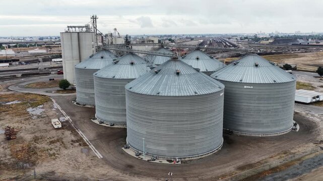 Cluster of steel grain silos rises at industrial storage facility near Fresno, California, USA, surrounded by conveyor towers, gravel yard under thick overcast cloudy sky. Aerial drone view. Wide shot
