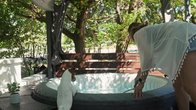 Woman cleaning an inflatable hot tub with a pool skimmer while a small dog watches beside the spa. Outdoor pool maintenance and summer backyard care.