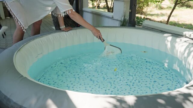 Woman cleaning leaves from an inflatable hot tub, using a pool skimmer to collect floating debris from the water. Outdoor spa cleaning and maintenance.