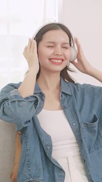 Happy Asian woman relaxing on sofa and listening to music with headphones, enjoying favorite song at home.