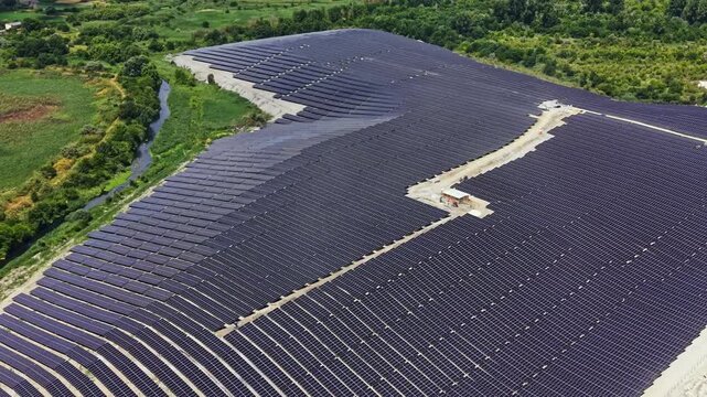 Electricity is generated by many solar panels lined up in neat rows. This facility is located near a natural landscape with trees and water. The sun is shining brightly over the scene.