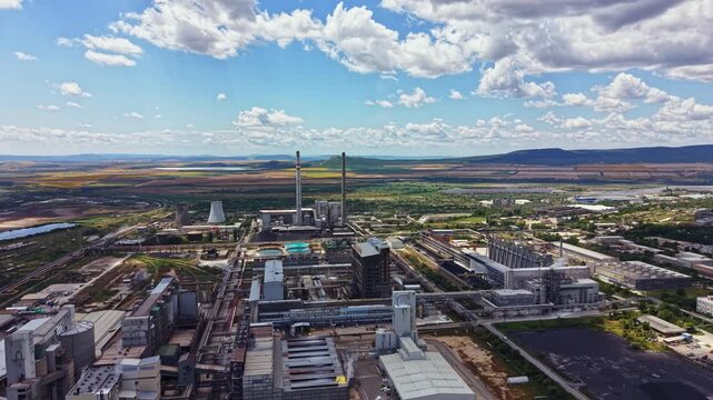 The scene shows a wide view of an industrial area featuring several large buildings and smokestacks. Green fields stretch out in the background under a clear blue sky with fluffy clouds.