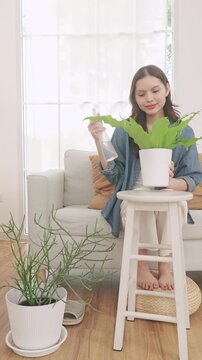 A young Asian woman sitting in her living room, using a spray bottle to mist water on a potted fern.