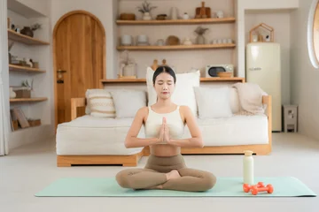 Asian woman in beige activewear sits in lotus pose with prayer hands and closed eyes during a calm home yoga session. © Jestercine