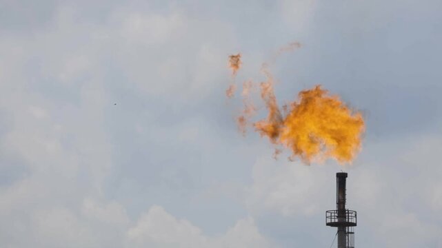 Industrial gas flare stack with burning flame against cloudy blue sky at oil and gas refinery
