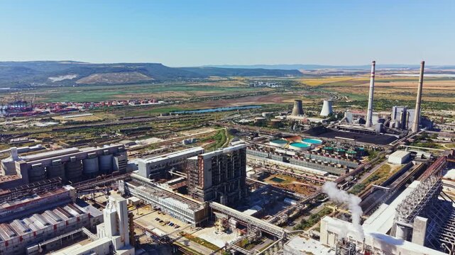 A large industrial complex is shown with various buildings and smoke stacks. The land around is flat and has fields along with some green areas. The sky is clear and blue.