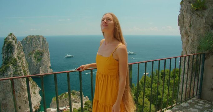 Smiling woman in yellow dress walkig on Capri balcony enjoying sunny sea view during tourist tour