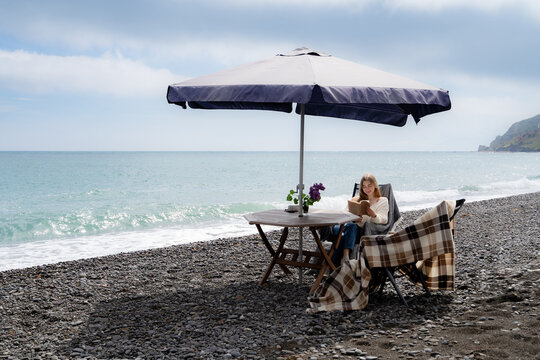 A woman is reading a book at a table on a pebbled beach by the sea, during a relaxing day outdoors.