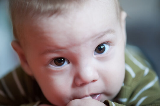 An extreme closeup of a sweet baby's face, displaying wide brown eyes full of curiosity. The infant gently sucks on a finger, absorbed in exploring sensations within a peaceful home setting